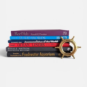 May include: A stack of six hardcover books titled Reef Fish, Island Worlds, Anemonefishes of the World, Ocean Liners of the 20th Century, Whale Nation, and Complete Encyclopedia of the Freshwater Aquarium, with a gold-toned metal ship wheel.
