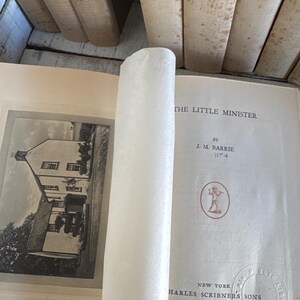 Puede incluir: Un libro abierto titulado "THE LITTLE MINISTER" de J.M. Barrie, publicado por Charles Scribner's Sons en Nueva York, MCMXXIX. Las p&aacute;ginas del libro est&aacute;n envejecidas, con una imagen en blanco y negro de un edificio y un papel blanco enrollado.