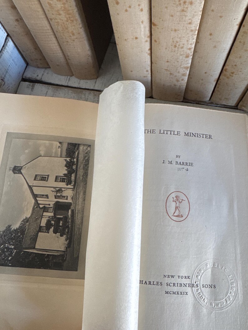 Puede incluir: Un libro abierto titulado "THE LITTLE MINISTER" de J.M. Barrie, publicado por Charles Scribner's Sons en Nueva York, MCMXXIX. Las p&aacute;ginas del libro est&aacute;n envejecidas, con una imagen en blanco y negro de un edificio y un papel blanco enrollado.