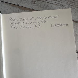 May include: A handwritten inscription on a white page. The text reads "MARTHA E. ANDERSON 409 CHICORY PL. FRUIT COVE, FL. 6/24/2002". The page is set against a light-colored, textured wooden surface.