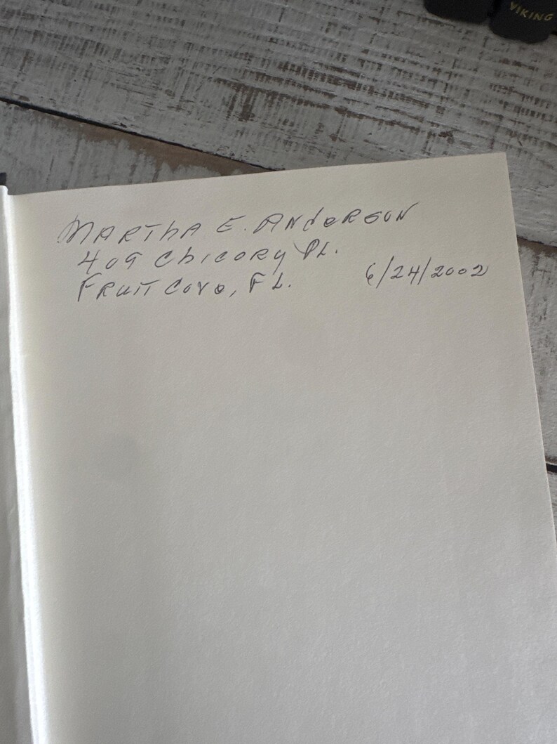 May include: A handwritten inscription on a white page. The text reads "MARTHA E. ANDERSON 409 CHICORY PL. FRUIT COVE, FL. 6/24/2002". The page is set against a light-colored, textured wooden surface.