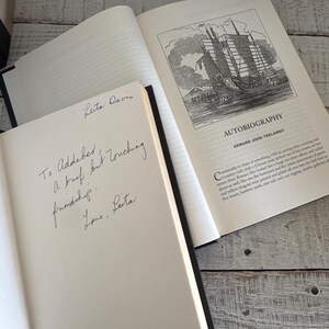 Pu&ograve; includere: Due libri aperti con dorsi neri su una superficie di legno chiaro. Un libro mostra un'illustrazione dettagliata di una nave e il titolo "AUTOBIOGRAPHY". L'altro libro ha un testo scritto a mano che recita "To Addeber, A brief but touching friendship. Love, Leita."