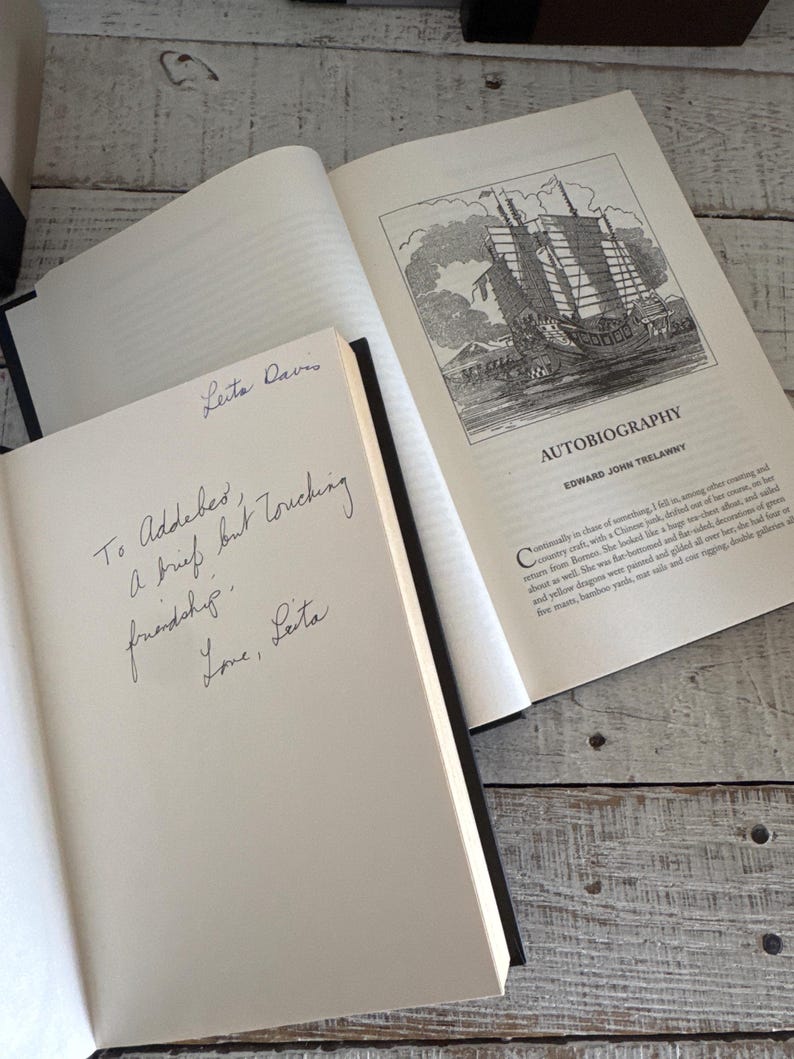 Pu&ograve; includere: Due libri aperti con dorsi neri su una superficie di legno chiaro. Un libro mostra un'illustrazione dettagliata di una nave e il titolo "AUTOBIOGRAPHY". L'altro libro ha un testo scritto a mano che recita "To Addeber, A brief but touching friendship. Love, Leita."