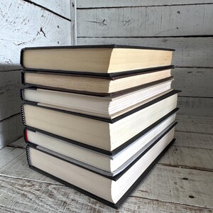 Pu&ograve; includere: Una pila di libri con copertina rigida, con dorsi neri e pagine color crema. I libri sono impilati ordinatamente, con le pagine leggermente ingiallite. Lo sfondo &egrave; una superficie di legno verniciata di bianco.