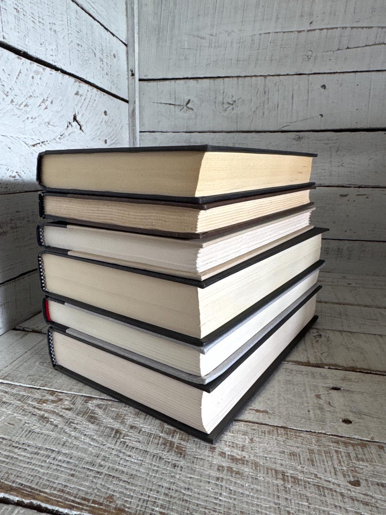 Pu&ograve; includere: Una pila di libri con copertina rigida, con dorsi neri e pagine color crema. I libri sono impilati ordinatamente, con le pagine leggermente ingiallite. Lo sfondo &egrave; una superficie di legno verniciata di bianco.