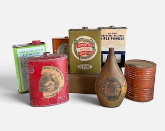 May include: A collection of vintage metal gunpowder and rifle powder containers. The group includes a red Shooting Gunpowder Co. tin, a green DuPont No. 1 Rifle Smokeless tin, a white and blue Du Pont Improved Military Rifle Powder tin, and a brown flask.