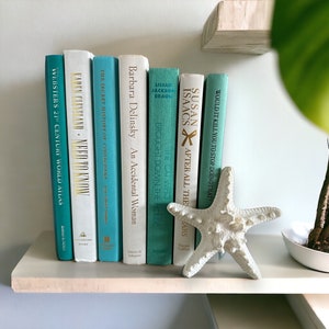 May include: A bookshelf with a white background and a white starfish on the shelf. The bookshelf is filled with teal and white books with gold lettering. The titles of the books are "Webster's 21st Century World Atlas", "Karen Cleveland - Need to Know", "The Secret History of Costachana", "Barbara Delinsky - An Accidental Woman", "The Cai Who Brought Down the House", "Susan Isaacs - After All the Years", and "Would It Kill You to Stop Doing That?"