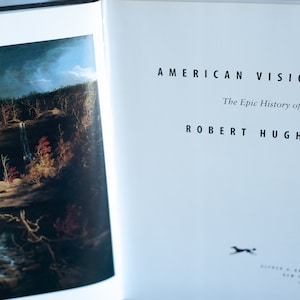 May include: Open book with the title "AMERICAN VISIONS" and the author "ROBERT HUGHES". The book's pages display a landscape painting with a waterfall and autumn foliage. The book is about the epic history of art in America.