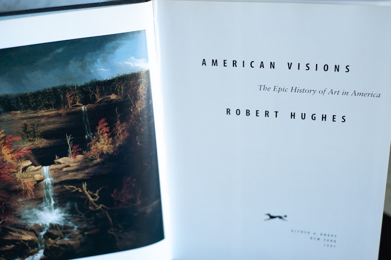 May include: Open book with the title "AMERICAN VISIONS" and the author "ROBERT HUGHES". The book's pages display a landscape painting with a waterfall and autumn foliage. The book is about the epic history of art in America.