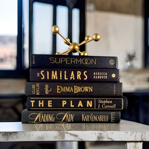 Puede incluir: Una pila de cinco libros de tapa dura negros con letras doradas sobre una superficie blanca. Los t&iacute;tulos de los libros son "Supermoon", "The Similars", "Emma Brown", "The Plan", y "Leading Lady". Un soporte decorativo de metal dorado con una parte superior redonda y tres patas est&aacute; encima de los libros.