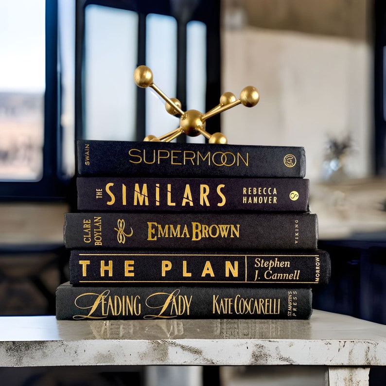Puede incluir: Una pila de cinco libros de tapa dura negros con letras doradas sobre una superficie blanca. Los t&iacute;tulos de los libros son "Supermoon", "The Similars", "Emma Brown", "The Plan", y "Leading Lady". Un soporte decorativo de metal dorado con una parte superior redonda y tres patas est&aacute; encima de los libros.