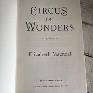 May include: Open book with the title "Circus of Wonders" by Elizabeth Macneal. The book's cover features elegant typography and the publisher's name, Emily Bestler Books, is visible.