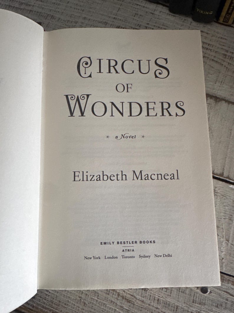 May include: Open book with the title "Circus of Wonders" by Elizabeth Macneal. The book's cover features elegant typography and the publisher's name, Emily Bestler Books, is visible.