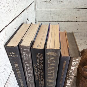 May include: A selection of antique books with dark grey covers and gold lettering. Titles include "The Secret Pilgrim," "Goddess," and "Majesty." The books are arranged on a white wooden surface, creating a classic aesthetic.