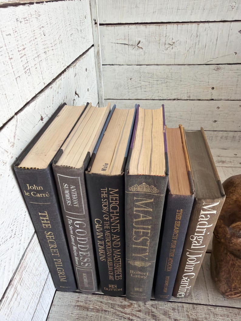 May include: A selection of antique books with dark grey covers and gold lettering. Titles include "The Secret Pilgrim," "Goddess," and "Majesty." The books are arranged on a white wooden surface, creating a classic aesthetic.