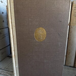Puede incluir: Un libro antiguo de tapa dura con una cubierta marr&oacute;n y un emblema dorado. El lomo es blanquecino y el libro muestra signos de edad. El libro forma parte de una colecci&oacute;n de libros antiguos.