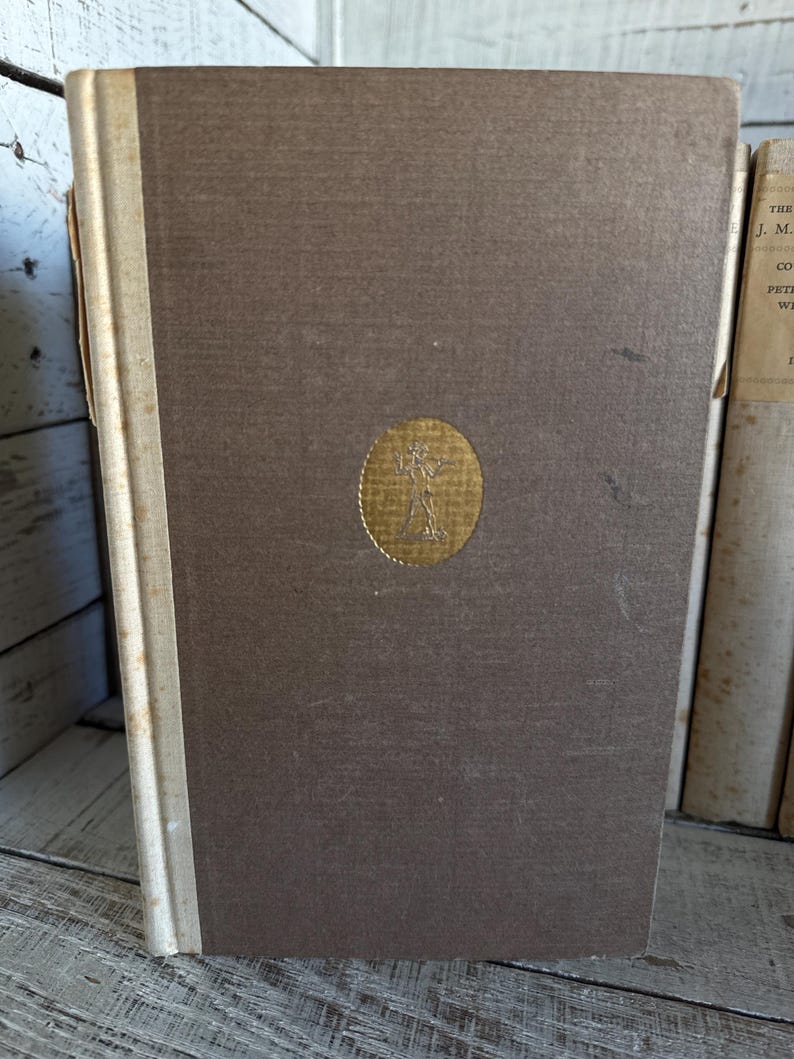 Puede incluir: Un libro antiguo de tapa dura con una cubierta marr&oacute;n y un emblema dorado. El lomo es blanquecino y el libro muestra signos de edad. El libro forma parte de una colecci&oacute;n de libros antiguos.