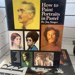 K&ouml;nnte beinhalten: Ein Buch mit dem Titel "Wie man Portr&auml;ts in Pastell malt" von Joe Singer. Der Umschlag zeigt eine Auswahl an Pastellportr&auml;ts von Menschen, darunter ein Mann mit Schnurrbart, eine Frau mit Brille und ein kleiner Junge.