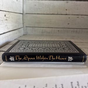 May include: A black hardback book with a silver and black patterned cover. The title "The Space Within The Heart" is written in gold lettering on the spine.