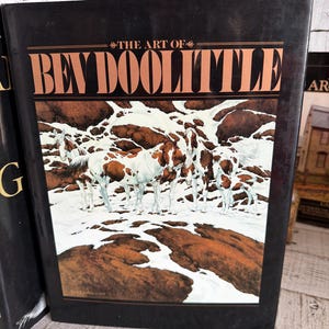May include: A hardcover book titled The Art of Bev Doolittle with a black cover featuring a painting of several brown and white horses standing in a snowy landscape with dark brown rock formations visible throughout the scene.