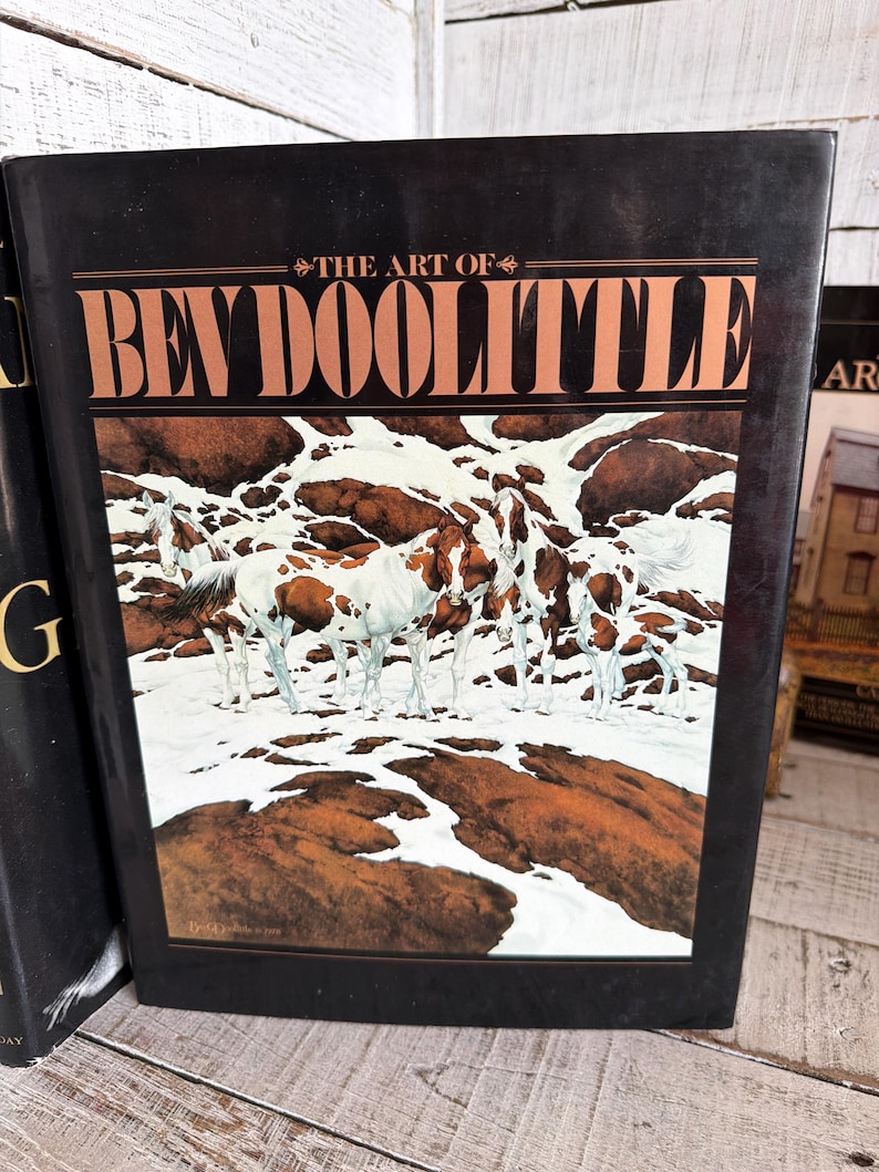May include: A hardcover book titled The Art of Bev Doolittle with a black cover featuring a painting of several brown and white horses standing in a snowy landscape with dark brown rock formations visible throughout the scene.