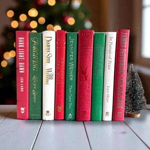 May include: A collection of hardcover books with red, green, and white spines. Titles include "Dark Light: Dawn," "Forgiving," "The Wedding," "Then Came You," and "The Red Book." A small decorative Christmas tree is on the right.