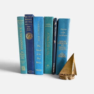 May include: A collection of six books with blue and teal spines standing upright, featuring gold lettering and titles including S.S. Savannah, The Use of Radar at Sea, and Great Voyages in Small Boats, accompanied by a small brass sailboat figurine.