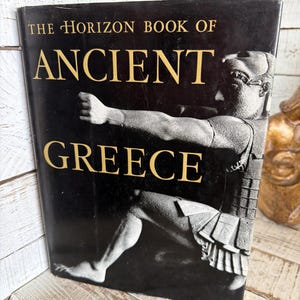 May include: A hardcover book titled The Horizon Book of Ancient Greece with a black dust jacket. The cover features gold lettering and a monochromatic photographic illustration of a classical Greek statue depicting a figure in a dynamic pose.
