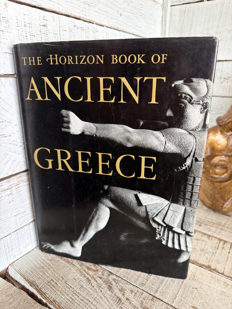 May include: A hardcover book titled The Horizon Book of Ancient Greece with a black dust jacket. The cover features gold lettering and a monochromatic photographic illustration of a classical Greek statue depicting a figure in a dynamic pose.
