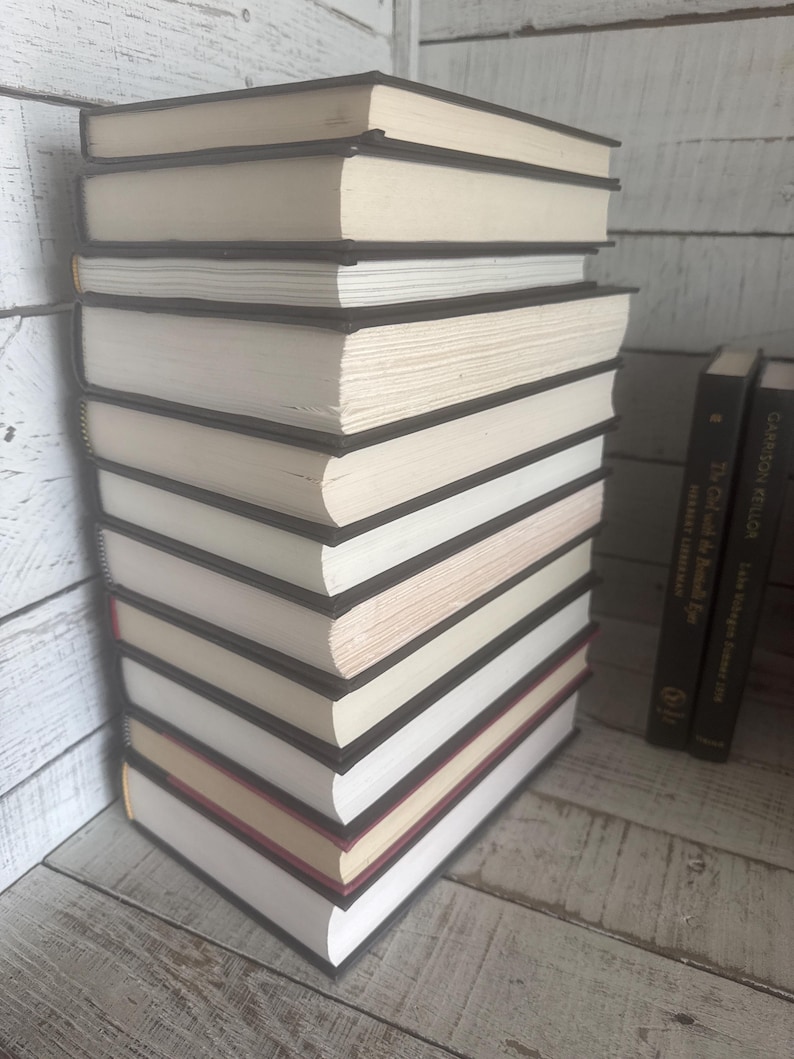 May include: A stack of hardcover books with black spines and off-white pages. The books are neatly arranged, with some showing red and gold accents on their edges. The background features a white wooden surface.
