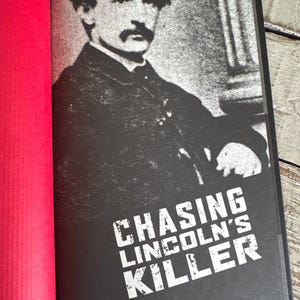May include: A book with a black cover featuring a grayscale image of a man in a hat and coat. The title "CHASING LINCOLN'S KILLER" is printed in white, bold letters. The book is open, revealing a red page.