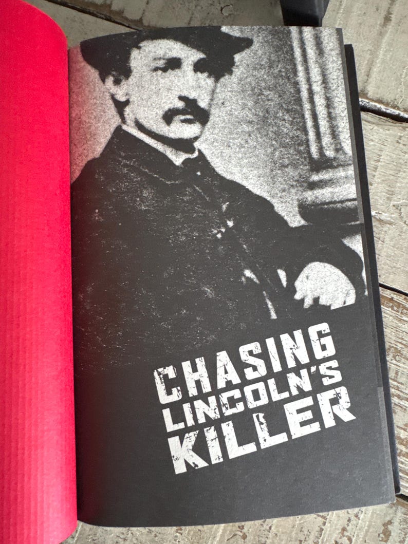 May include: A book with a black cover featuring a grayscale image of a man in a hat and coat. The title "CHASING LINCOLN'S KILLER" is printed in white, bold letters. The book is open, revealing a red page.