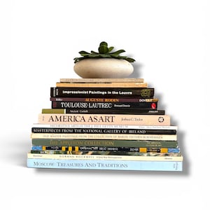 May include: A stack of books with titles like "America as Art" and "Moscow: Treasures and Traditions." A small, light-colored stone with a green plant on top sits atop the stack. The books are various colors.