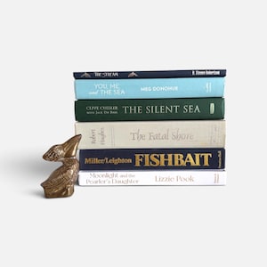 May include: A stack of six hardcover books with a bronze-colored metal pelican figurine placed to the left. The book titles include The Stream, You, Me, and the Sea, The Silent Sea, The Fatal Shore, Fishbait, and Moonlight and the Pearler's Daughter.