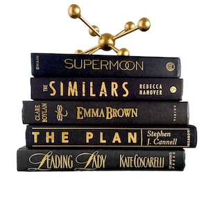 Puede incluir: Una pila de cinco libros de tapa dura negros con letras doradas en los lomos. Los t&iacute;tulos son "Supermoon", "The Similars", "& Emma Brown", "The Plan", y "Leading Lady". Un soporte decorativo de metal dorado con una parte superior redonda y tres patas se coloca encima de los libros.