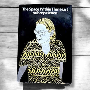 May include: A black book cover with the title "The Space Within The Heart" and the author "Aubrey Menen". The cover features a black and white illustration of a person wearing a yellow and black patterned shirt.