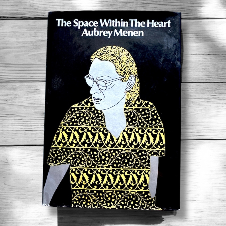 May include: A black book cover with the title "The Space Within The Heart" and the author "Aubrey Menen". The cover features a black and white illustration of a person wearing a yellow and black patterned shirt.