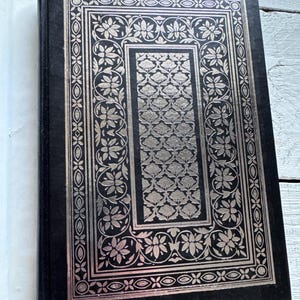 May include: A black hardback book with a silver embossed design on the front cover. The design features a floral pattern with intricate details.