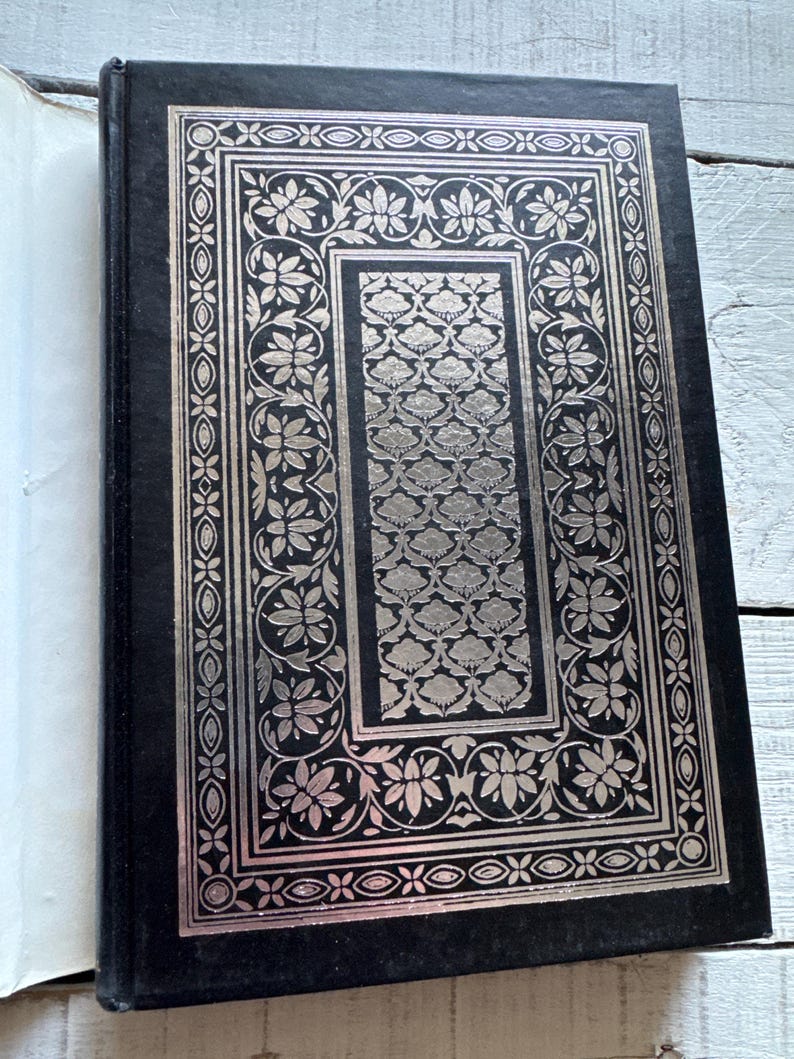 May include: A black hardback book with a silver embossed design on the front cover. The design features a floral pattern with intricate details.