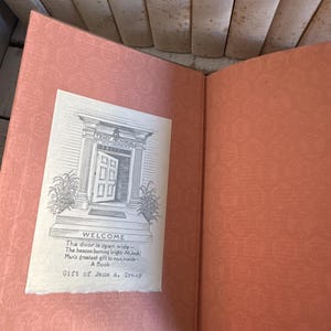 Puede incluir: Libro abierto con una cubierta y p&aacute;ginas de color melocot&oacute;n. Una ilustraci&oacute;n en blanco y negro de la entrada de un edificio con la palabra "WELCOME" est&aacute; en un papel blanco. El texto debajo de la ilustraci&oacute;n dice "Gift of Jane A. Troy."
