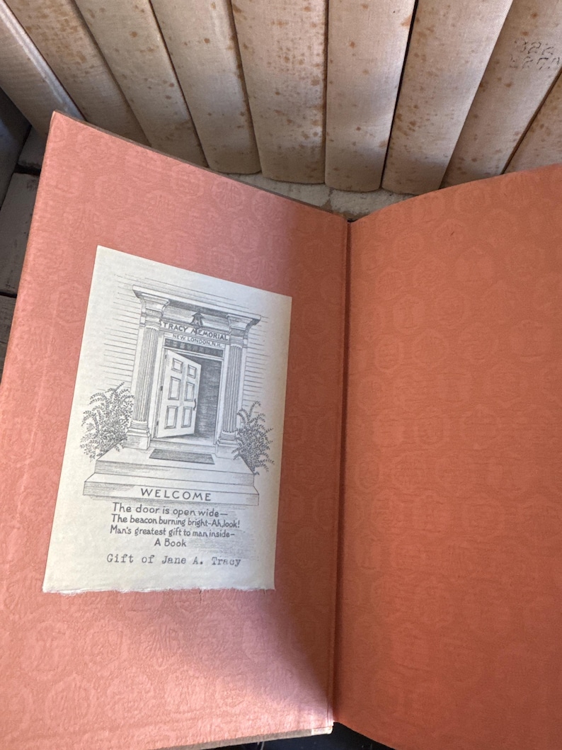 Puede incluir: Libro abierto con una cubierta y p&aacute;ginas de color melocot&oacute;n. Una ilustraci&oacute;n en blanco y negro de la entrada de un edificio con la palabra "WELCOME" est&aacute; en un papel blanco. El texto debajo de la ilustraci&oacute;n dice "Gift of Jane A. Troy."