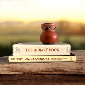 May include: A small, brown ceramic vase with a black and white geometric design sits atop two books. The top book is titled "The Indians' Book" and the bottom book is titled "The North American Indians". The books are stacked on a wooden surface.