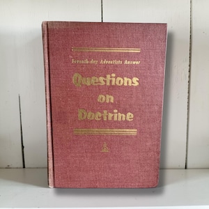 Może przedstawiać: Vintage książka z teksturowaną, czerwonobrązową okładką. Złoty napis głosi "Seventh-day Adventists Answer Questions on Doctrine". Książka ma mały emblemat na dole. Książka stoi pionowo.