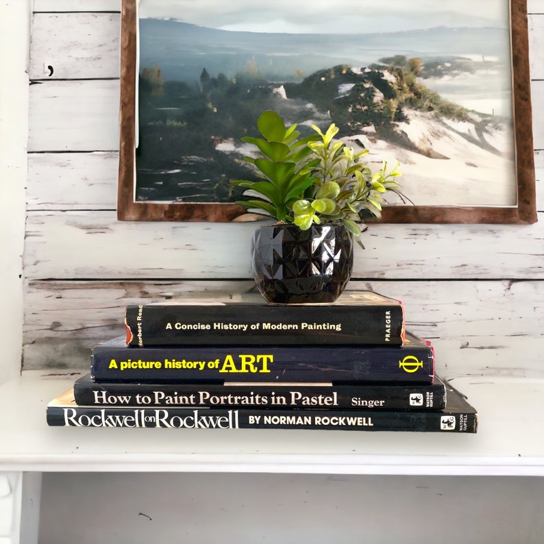 K&ouml;nnte beinhalten: Ein Stapel von vier schwarzen Hardcover-B&uuml;chern mit gelben Texttiteln. Das oberste Buch tr&auml;gt den Titel "Rockwell on Rockwell" von Norman Rockwell. Das zweite Buch tr&auml;gt den Titel "How to Paint Portraits in Pastel". Das dritte Buch tr&auml;gt den Titel "A picture history of ART". Das unterste Buch tr&auml;gt den Titel "A Concise History of Modern Painting". Eine kleine Topfpflanze mit gr&uuml;nen Bl&auml;ttern steht auf den B&uuml;chern.