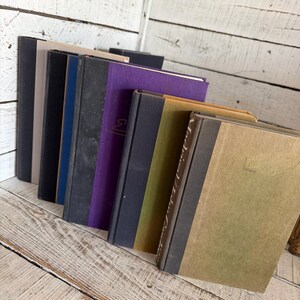 May include: A selection of antique books with diverse coloured covers, including black, navy blue, grey, and purple. The books are arranged on a distressed, white-painted wooden surface, creating a rustic aesthetic. Some books have gold lettering.