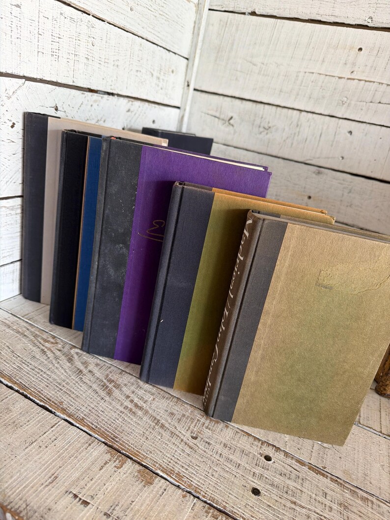 May include: A selection of antique books with diverse coloured covers, including black, navy blue, grey, and purple. The books are arranged on a distressed, white-painted wooden surface, creating a rustic aesthetic. Some books have gold lettering.