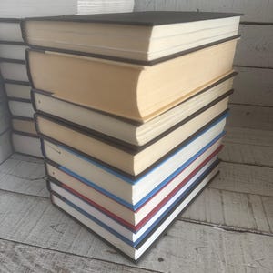 May include: A stack of hardcover books with black covers and cream-colored pages. The edges of the pages are painted in various colors, including red, white, and blue. Other books are stacked in the background.