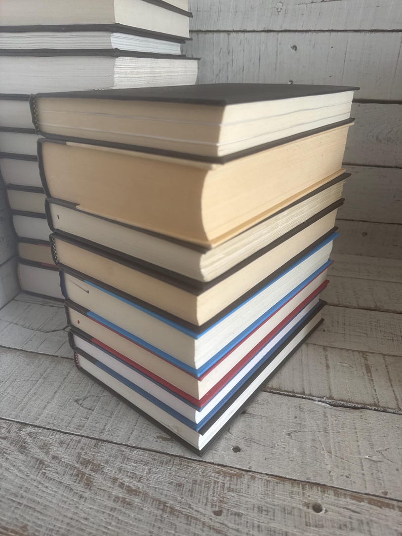 May include: A stack of hardcover books with black covers and cream-colored pages. The edges of the pages are painted in various colors, including red, white, and blue. Other books are stacked in the background.