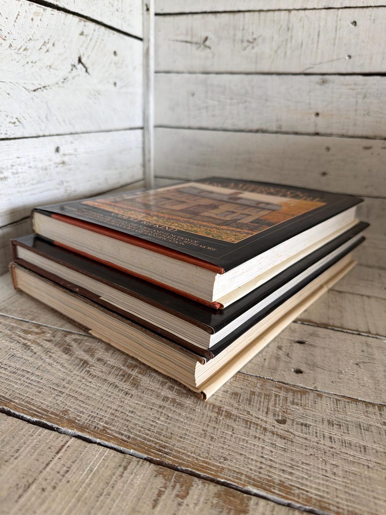 May include: A stack of three hardcover books with white pages. The top book features a dark cover with a rectangular image of a house and text that reads "CAROLE RIFKIND" and "A FIELD GUIDE TO AMERICAN ARCHITECTURE". The middle and bottom books have plain covers.