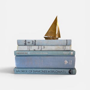May include: A stack of five light blue hardcover books with visible titles and author names, topped with a small brass sailboat figurine. The books are arranged horizontally, featuring text on the spines including "Baldrige of Diamonds & Diplomats".